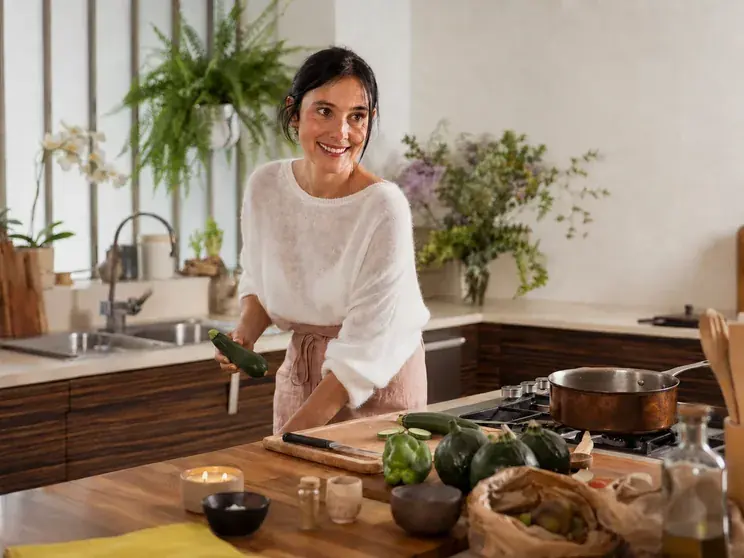 Mulher sorrindo enquanto cozinha legumes frescos em uma cozinha clara e acolhedora.