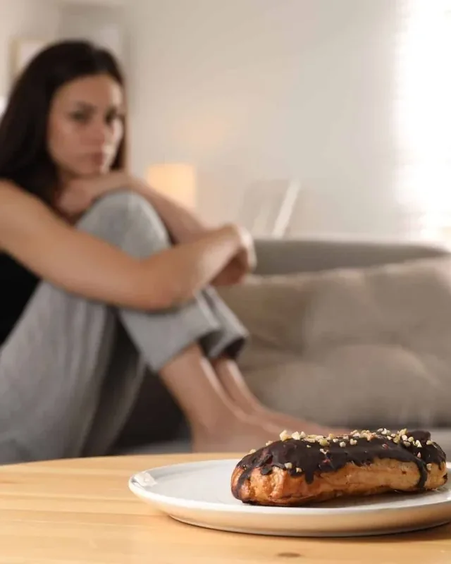 Mulher sentada ao fundo, pensativa, com um doce em primeiro plano sobre a mesa.